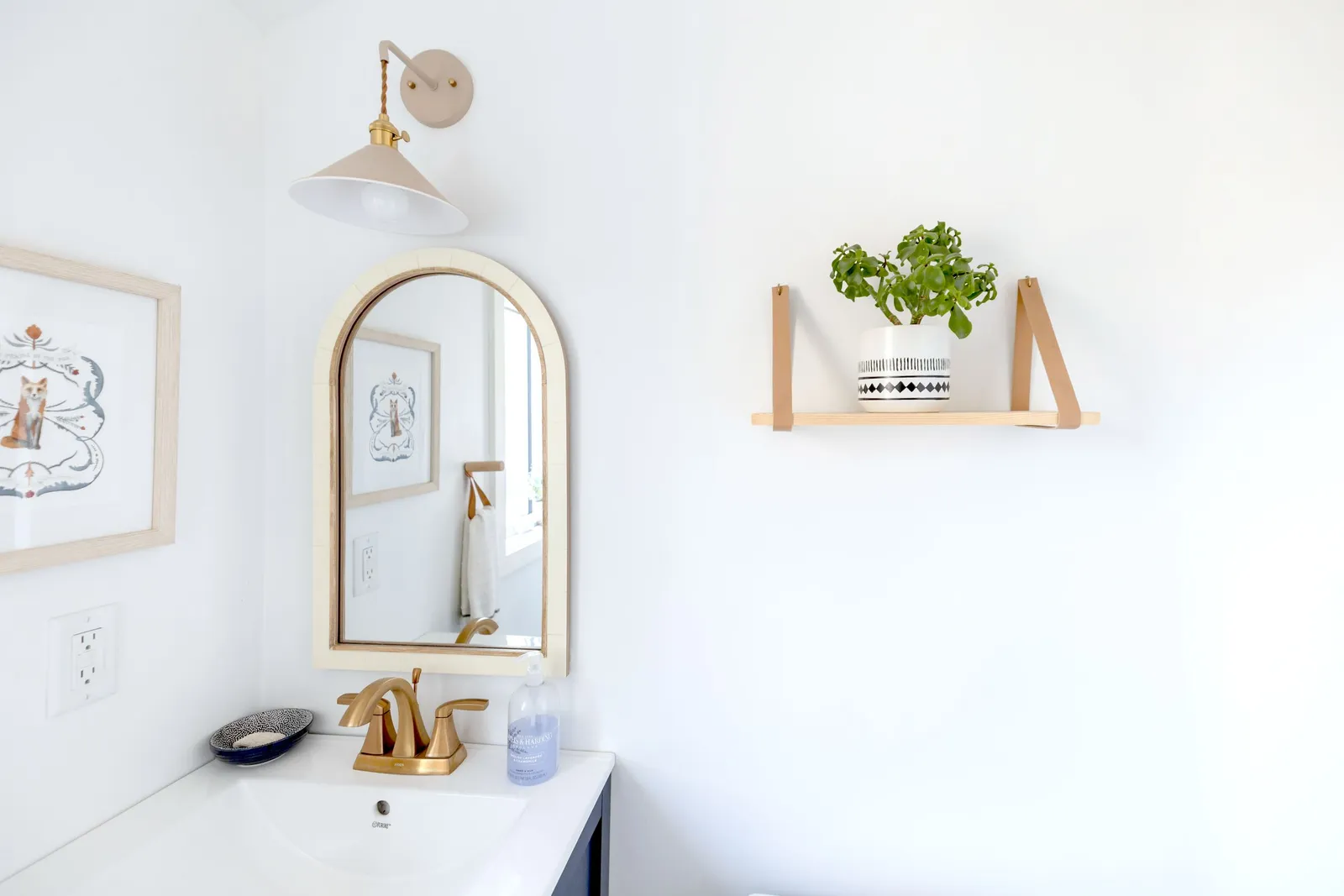 Small bathroom with a single vanity, a brass arched mirror, a wall shelf with a plant and a brass sconce